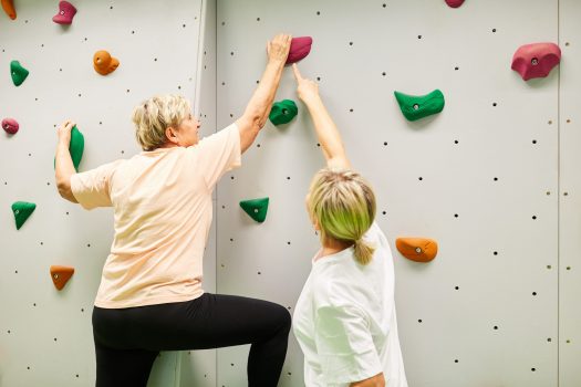 Vitale Seniorin beim Klettern an einer Kletterwand unter Anleitung einer Physiotherapeutin